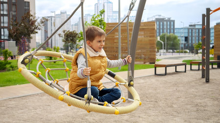 Little boy enjoying swinging in net swing on modern public playground at yard. Active child, sports and development, kids playing outdoors.の写真素材
