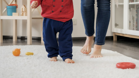 Closeup of little baby's feet learning walking with mother on soft carpet in living room. Baby...