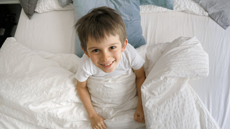 A young boy jumping out from under his blanket with a cheerful laugh and a look of playful happiness on his face looking in camera.の写真素材