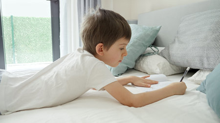 Cute boy doing his homework after waking up at morning and lying in bed. Concept of education, child...