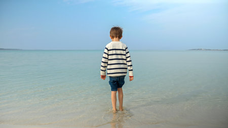 Rear view of little barefoot boy in sweater walking into the cold sea water on beach. Holiday,...