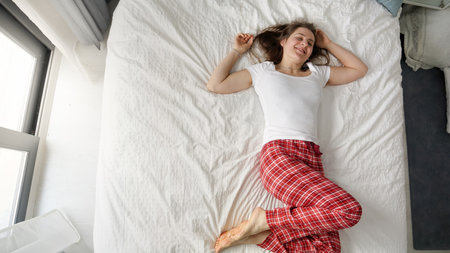 Young woman in pajamas lying on a bed with a big smile near a large window. Concept of weekend...