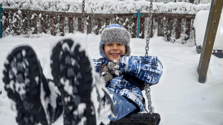 Cheerful laughing boy in warm hat and coat swinging on swing on playground covered with snow in winter.の写真素材