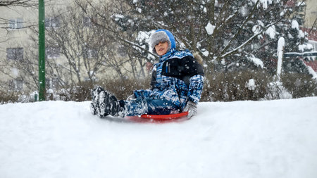 Cheerful smiling boy riding down the snowy hill on red plastic sleds during snowfall