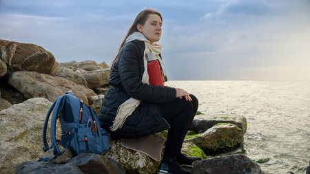 Young woman with long flowing hair sitting on a rocky coastline, holding a thermos of hot tea as she...