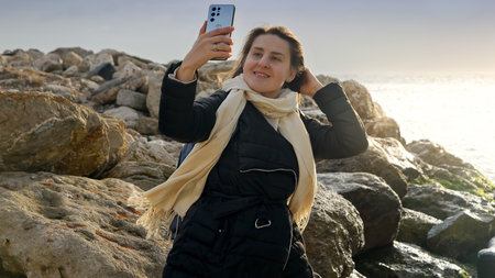 Young woman taking photos or herself at the winter sea using her smartphone while standing on the...
