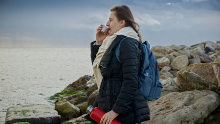 Young woman relaxing by the sea, holding a thermos of hot tea and admiring the wild and rugged coastline. Ideal for use in tourism, travel, or lifestyle contentの写真素材