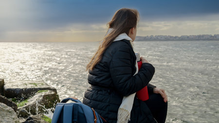 Young woman with long hair sitting on the big rocks at sea coast and looking on stormy weather over the ocean. Concept of tourism, travel, journey, holiday and vacation at winterの写真素材