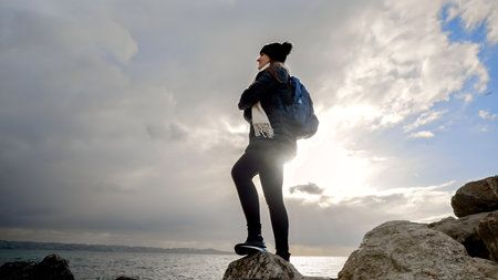 Young woman standing on top of a mountain, looking at the sunset over sea. With a backpack on her back, she takes in the beauty of nature. Concept of adventure, exploration, travel, and hikingの写真素材