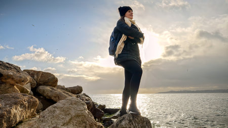 Young woman with backpack standing on cliff or rock top and looking at the sunset over sea on cold...