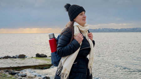 Smiling young woman with a backpack and thermos walking along a rocky sea beach, braving the cold and windy weather in pursuit of adventure and explorationの写真素材