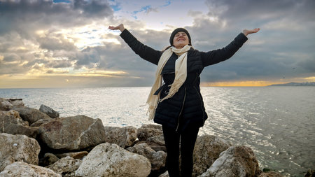Woman with a backpack stands on a sea cliff, arms stretched out, enjoying the magnificent view of...