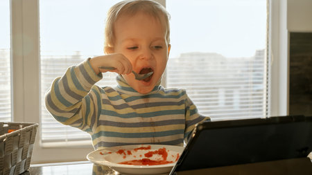 Little boy getting dirty and messy while eating soup on kitchen with tablet computer. Domestic food, gadget addiction, children healthy nutritionの写真素材