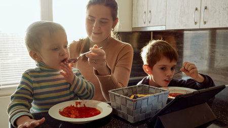 Young caring mother feeding her two sons with soup on the kitchen filled with sunset light.