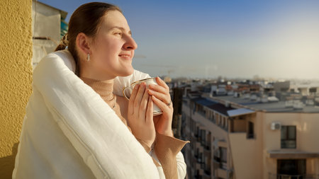 Smiling woman looking at sunset and drinking coffee on the balcony. People in morning, relaxing at home, enjoying city viewの写真素材