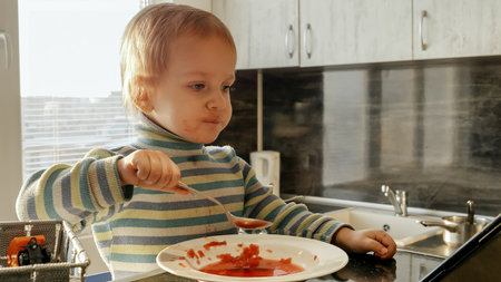 Cute messy boy eating soup with spoon and watching cartoons. Domestic food, gadget addiction, children healthy nutritionの写真素材