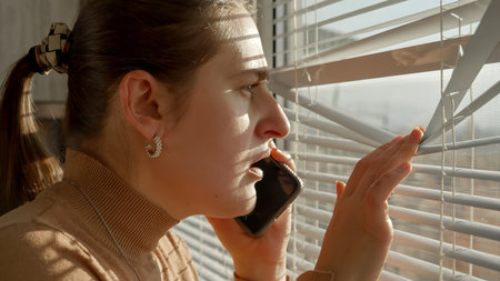 Portrait of stressed and scared woman loooking through window blinds on the street after seeing a crime.の写真素材