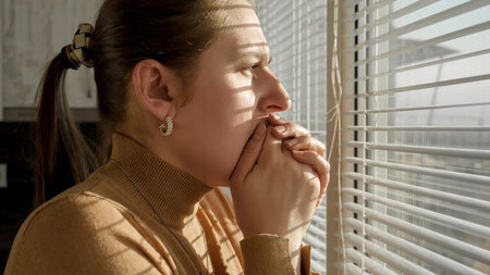 Afraid and scared woman closing her mouth with hand and looking out of the window through blinds.の写真素材
