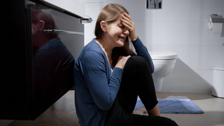 Crying woman sitting on floor in bathroom and leaning on sink.の写真素材