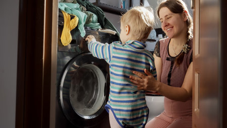 Little baby boy helping mother doing laundry in washing machine. Doing housework and chores, children education and developmentの写真素材