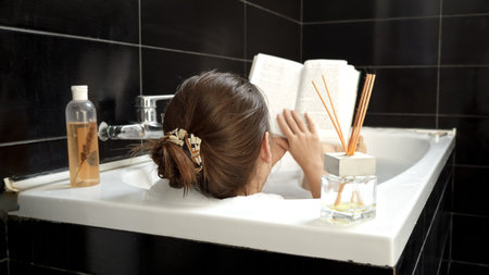 Rear view of young woman relaxing in bath and reading a book. Self-care, education, hygiene and spaの写真素材