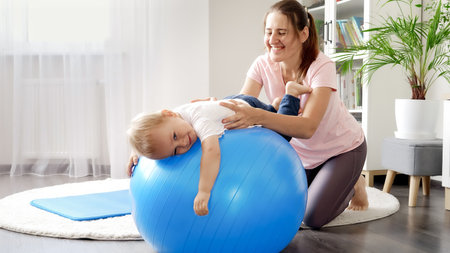 Smiling mother and baby doing fitness exercises on fitball at home. Concept of child healthcare, kids sports and yogaの写真素材