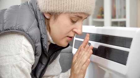 Woman in hat warming her hands at electric heater in cold house. Concept of energy crisis, high bills, economy and saving money on monthly utility paymentsの写真素材