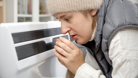 Portrait of stressed woman in warm hat feeling cold at home sitting at the heater.の写真素材