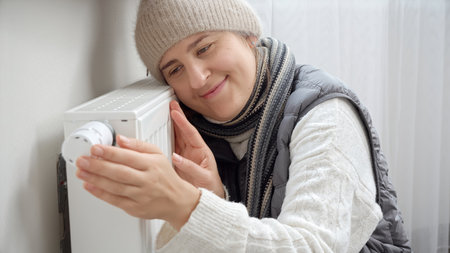 Portrait of smiling woman in warm clothes embracing heating radiator in cold house. Concept of energy crisis, high bills, broken heating system, economy and saving money on monthly utility paymentsの写真素材