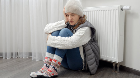 Young woman in warm clothes sitting at broken heating radiator in house. Concept of energy crisis, high bills, broken heating system, economy and saving money on monthly utility paymentsの写真素材