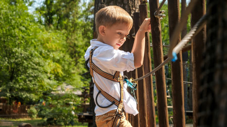 Portrait of young boy climbing and crossing rope bridge with wooden planks. Active childhood, healthy lifestyle, kids playing outdoors, children in natureの写真素材