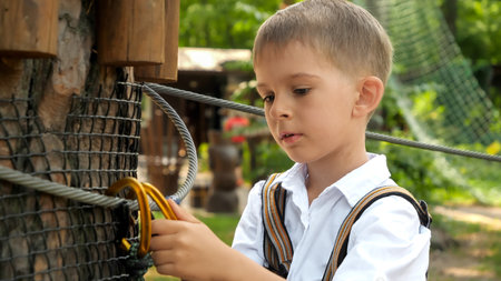 Little boy adjusting safety rope and hook in rope adventure park. Active childhood, healthy lifestyle, kids playing outdoors, children in natureの写真素材