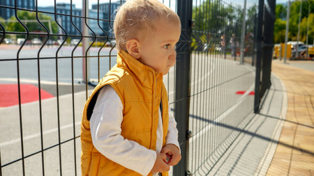 Little baby boy leaning on the metal net fence at playground. Upset children, negative emotions, kids problemsの写真素材