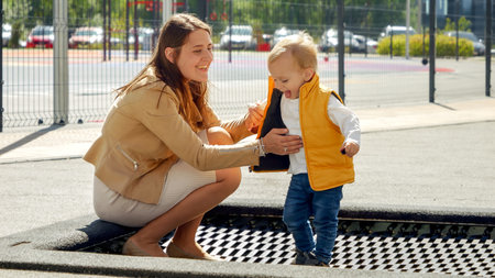 Cute baby boy holding mothers hand while jumping on trampoline at playground. Happy parenting, family having time together, kids and parents outdoorsの写真素材
