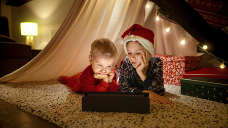 Two boys in pajamas lying under stairs at home and watching video on tablet computer while waiting for Christmas eve celebrations. Winter holidays, celebrations and partyの写真素材