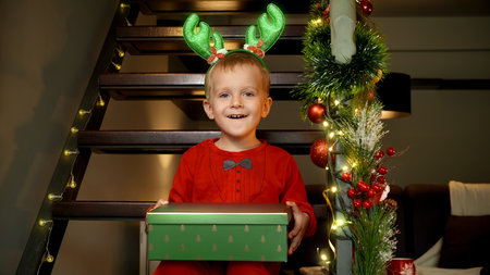 Portrait of cute toddler boy with funny antlers holding Christmas gift and sitting on wooden stairs at house on New Year celebration.の写真素材