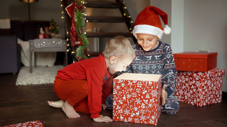 Two happy and excited boys in pajamas sitting on wooden floor and opening Christmas gifts and boxes with presents from Santa. Family celebrations on winter holidays.の写真素材