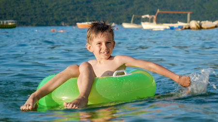 Happy cheerful boy splashing water while sitting in inflatable ring and swimming on calm sea waves at sunset. Family holidays, vacations, summertimeの写真素材