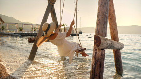 Young brunette woman in long dress swinging on rope swing at ocean beach bar. Tourism, summertime, holiday and vacation on beachの写真素材