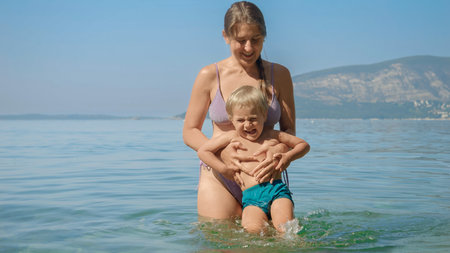 Cute smiling toddler boy having fun and splashing sea water while mother is holding himの写真素材