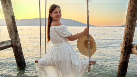 Young brunette woman in a flowing white dress, swinging by the sea beach in slow motion while enjoying the view of the setting sun. Travel, summertime bliss, holiday relaxation, and beachfront getaways.の写真素材