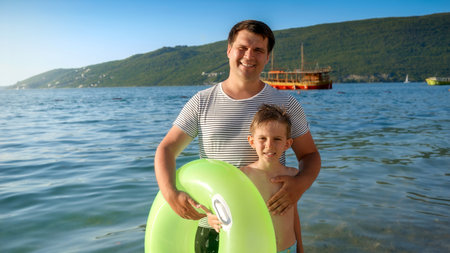 Happy smiling boy with young father standing on beach with inflatable ring before swimming in the sea. Family holidays, vacations, summertimeの写真素材