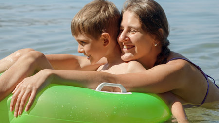 Portrait of happy cheerful mother swimming in sea with son sitting in inflatable ring.の写真素材
