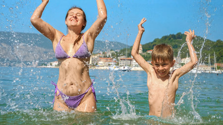 Happy cheerful excited boy splashing water with mother while relaxing on sunny sea beach.の写真素材