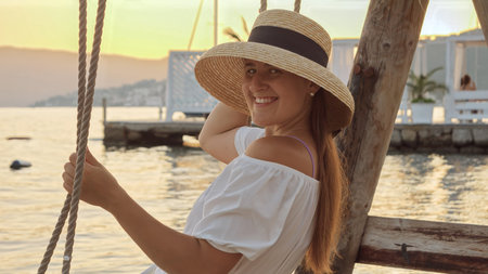 Portrait of happy smiling woman in straw hat sitting on the swing against sunset over the sea beach.の写真素材