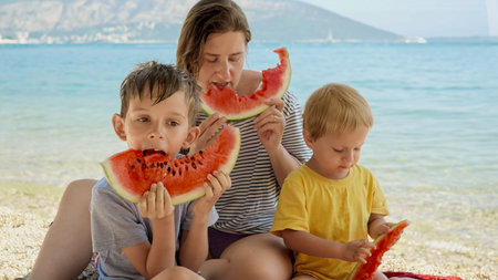 Happy mother with two boys sitting on the sea beach and enjoying eating sweet watermelon. Concept of family vacation, summertime, holiday and parents enjoying time with kidsの写真素材