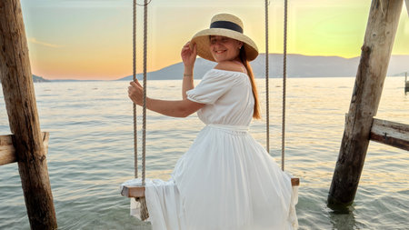 Happy smiling woman with long hair sitting on the swing at the sea and taking off straw hat.の写真素材