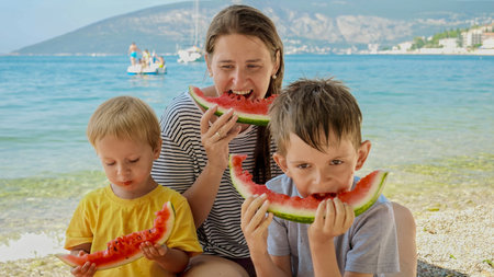 Happy mother and her two boys share laughter and sweet watermelon on a beachside picnic. Family vacation, the happiness of holidays, and parents relishing time with their children.の写真素材