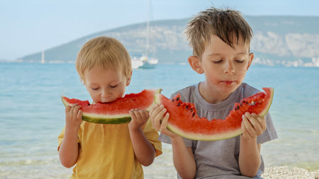 Two adorable boys eating sweet watermelon on a beach towel by the sea. Essence of summertime fun, holiday celebrations, and the relaxation of a sun-soaked vacation.の写真素材