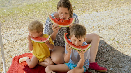 Happy mother and her two sons, sharing sweet watermelon on the sea beach. Summertime, holiday happiness, family vacation delight, and parents enjoying quality time with their kids.の写真素材